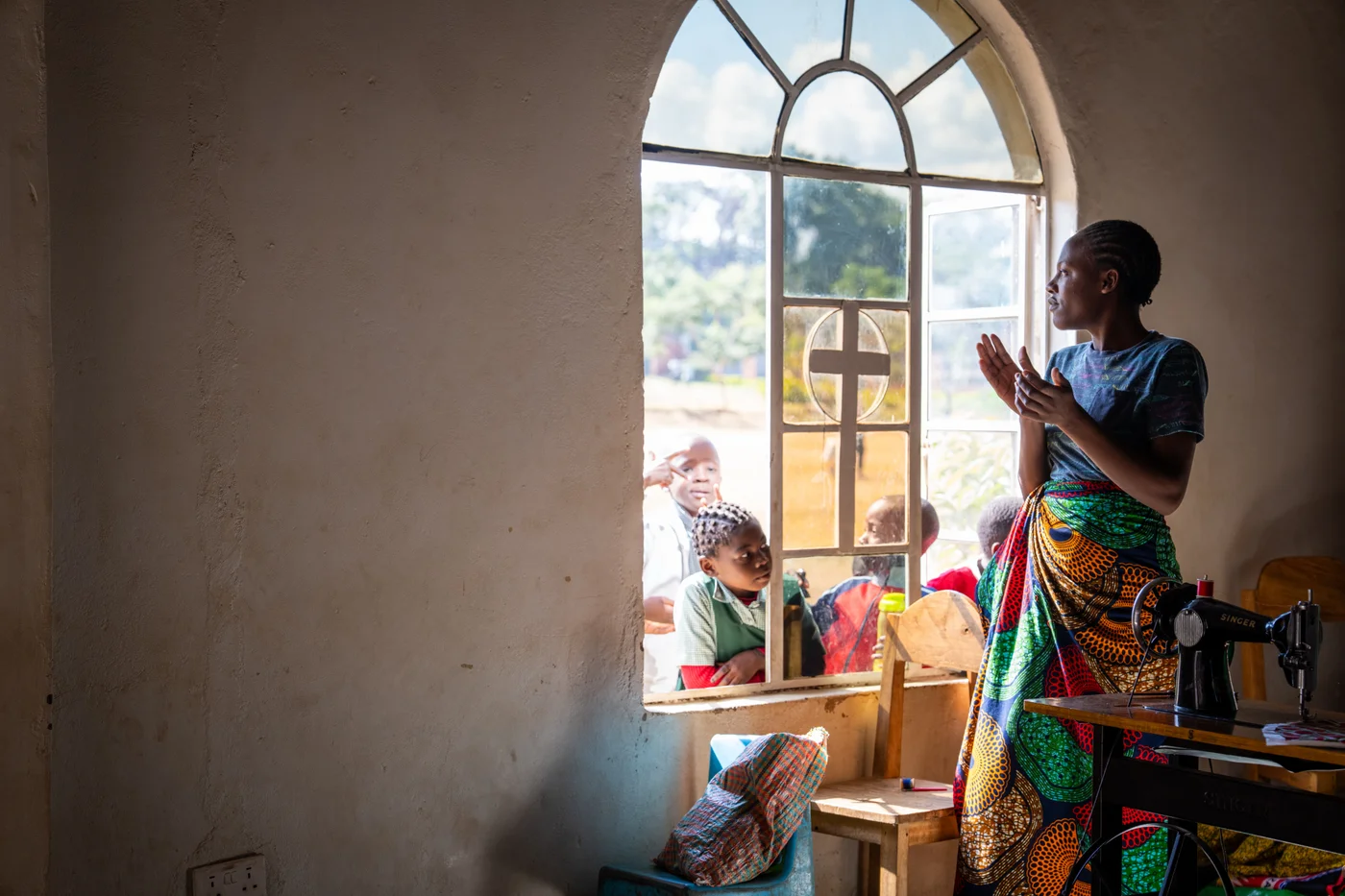 A woman beside a sewing machine and an arched window with children outside