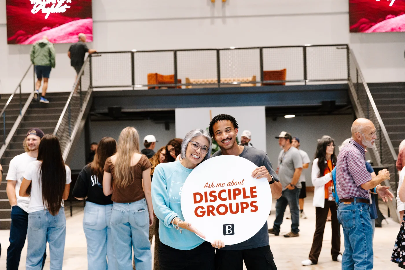 Two people holding an Ask me about Disciple Groups sign in a church lobby