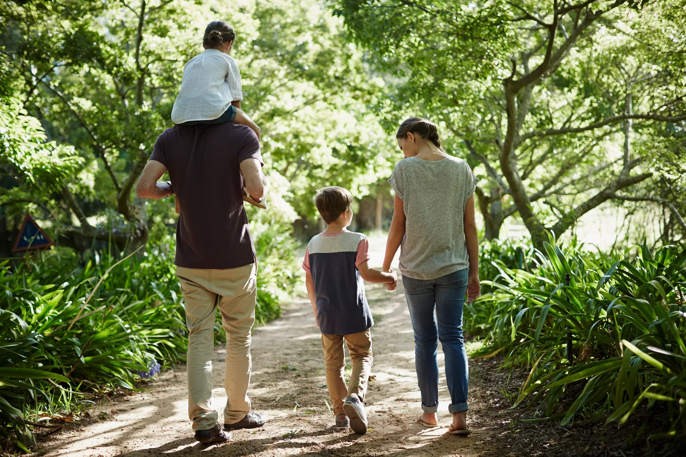 Two adults and two children walking along a leafy path, with one child riding on an adult’s shoulders
