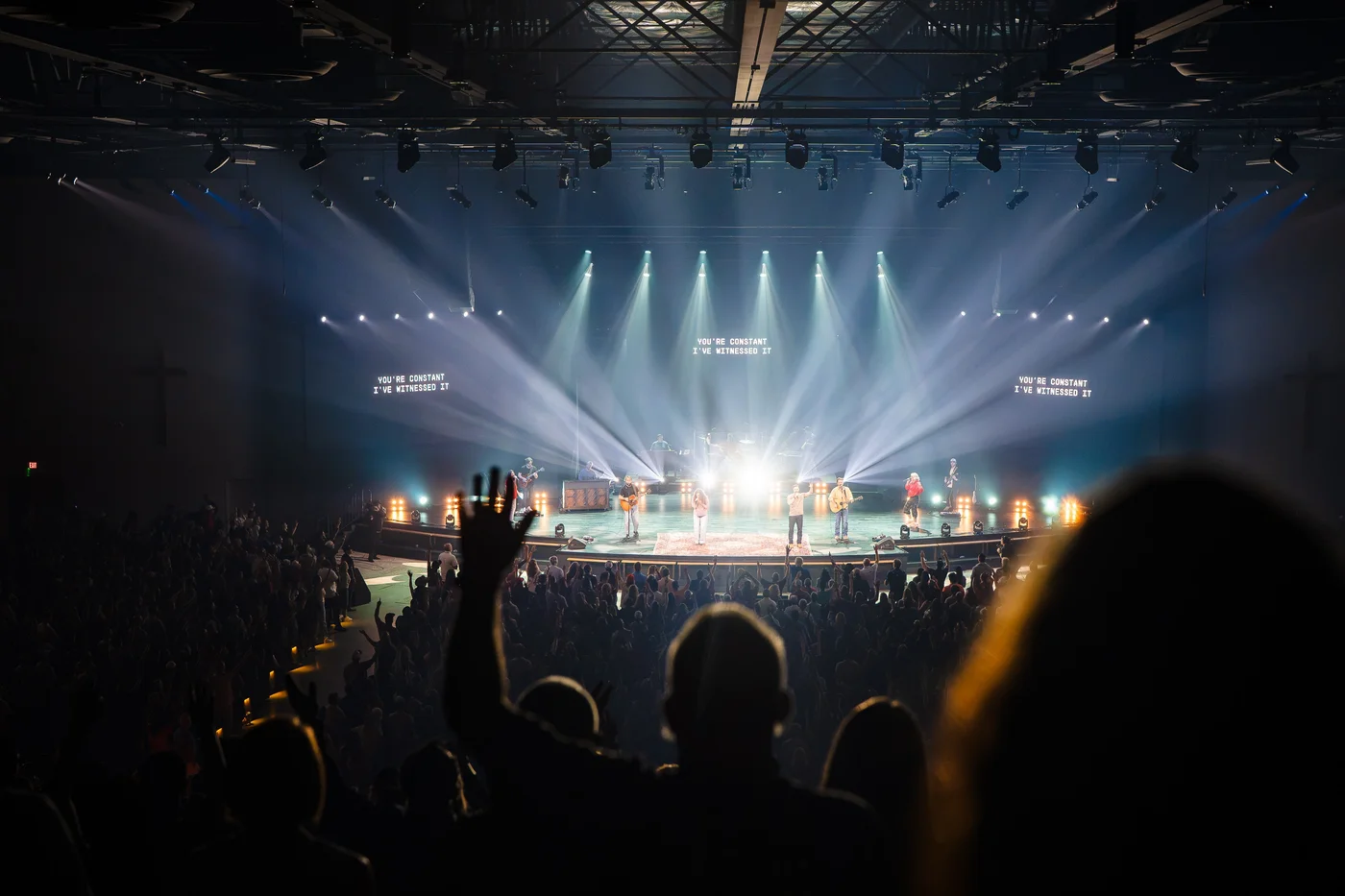 Conference attendees worshiping with hands raised toward a brightly lit stage